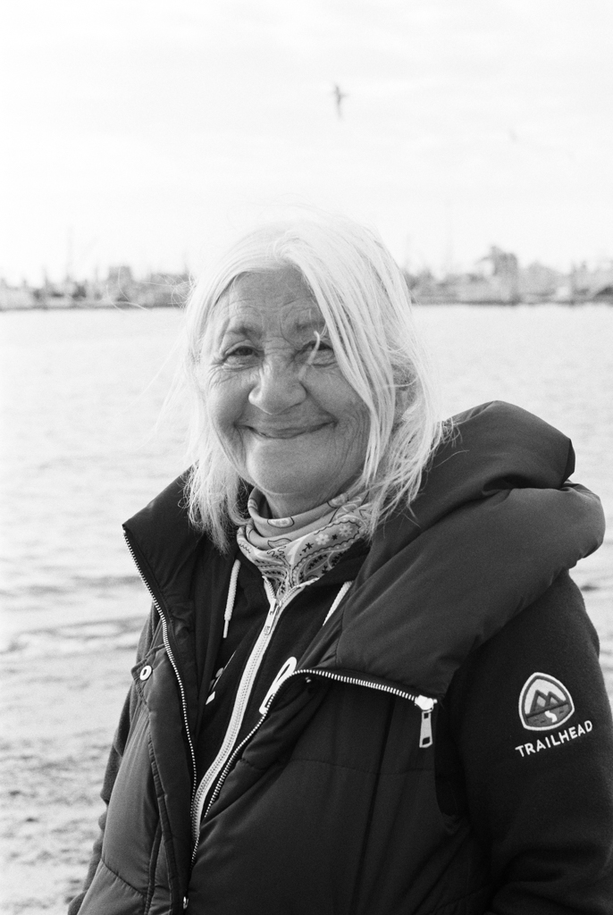 An elderly woman with silvery hair stands on the beach wearing a parka. A seagull flies by in the distance.