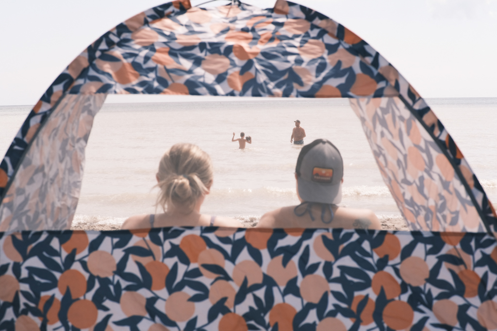 Through the back window of a beach tent, the heads of two lovers look out at the ocean. Between them, out in the water, a boy plays catch with his father.