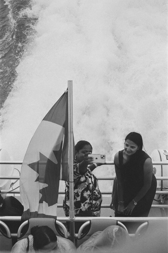 On the back of a ferry boat with the Canadian flag, two women lean on the rail and take a selfie with the wake in the background.