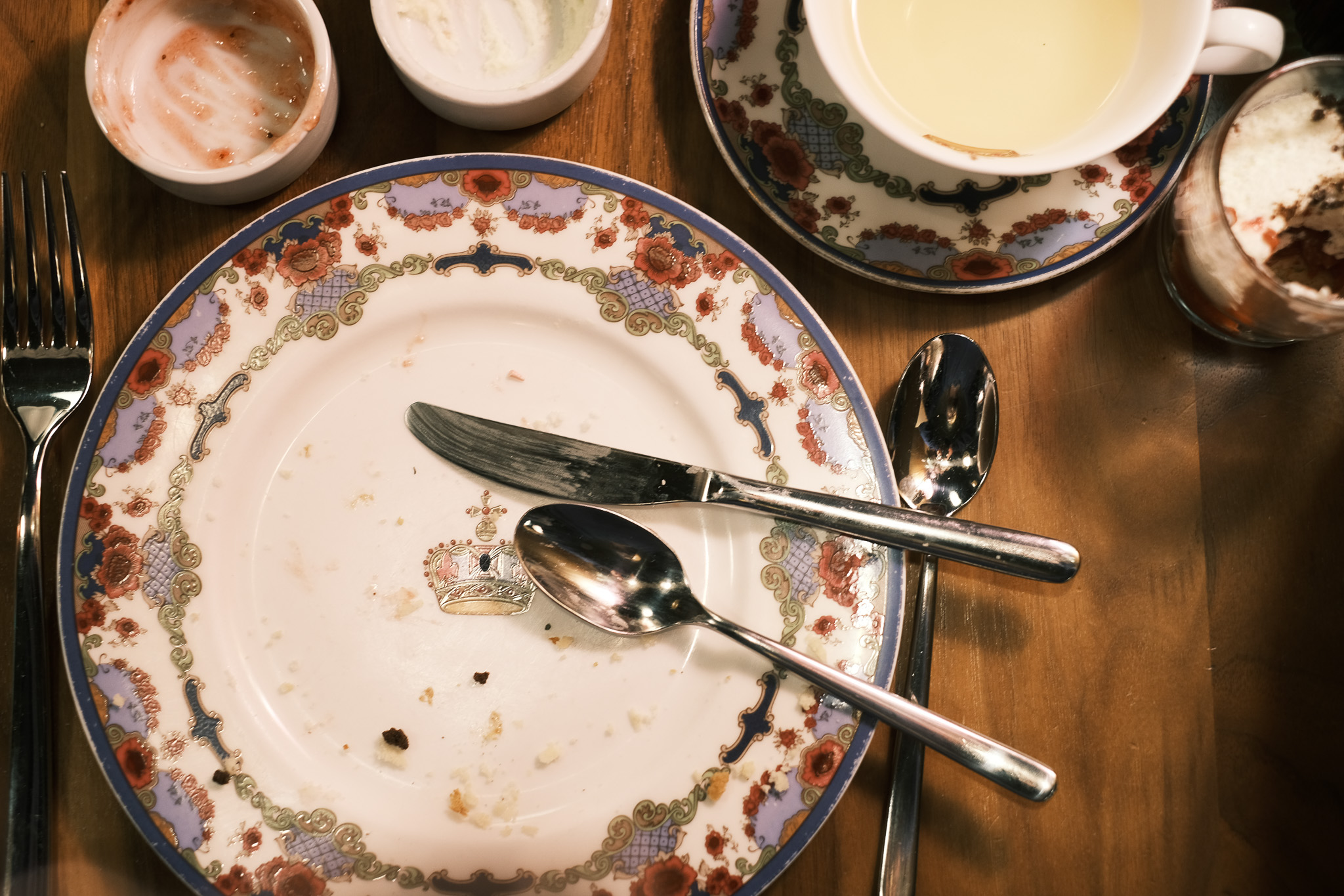 Looking down at a fancy china plate with the last crumbs of a delicious meal scattered upon it. A dirtied knife and spoon sit on the plate, and empty ramekins that once contained jelly and clotted cream are just on the edge of the frame. A half drunk cup of tea in the corner.
