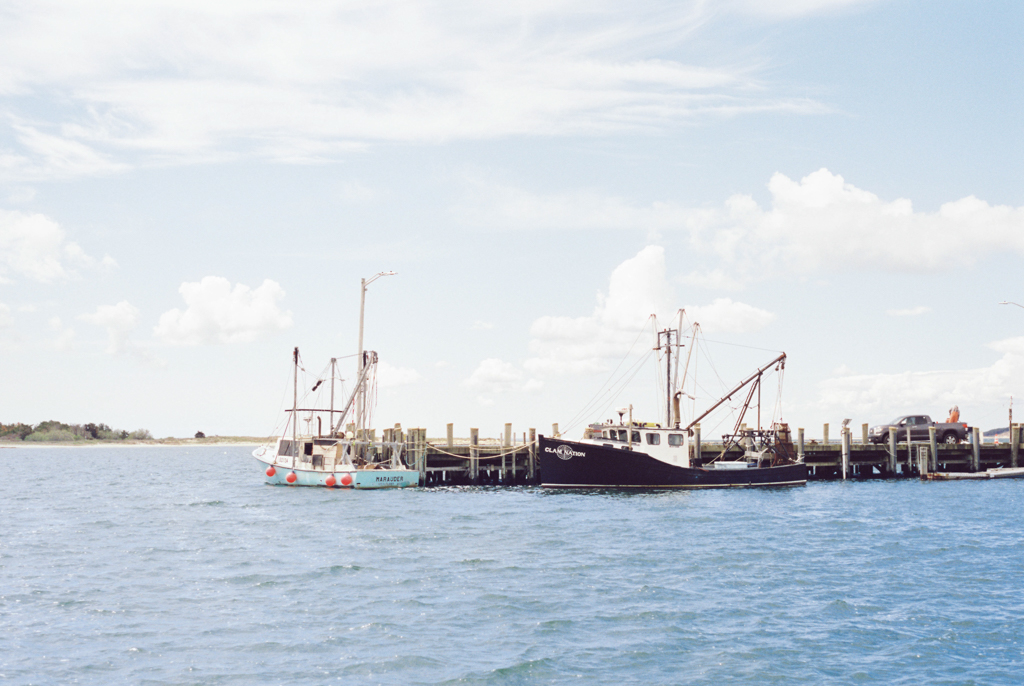 Boats docked in a cerulean blue bay, perfectly aligned with the horizon. On the far end of the dock, a man stands in the bed of his truck, shoveling lobster trap bait into a bucket.