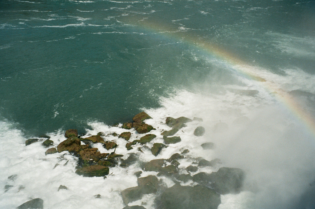 The tumult at the base of Niagara Falls creates white froth between the rocks and a rainbow in the mist.