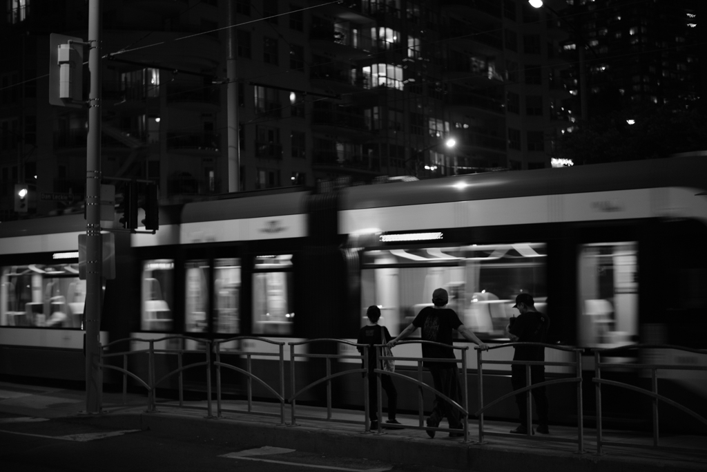 Three teens lean against the rail that separates the road from the light rail platform in Toronto's Harbourfront. The light rail zooms past them in a blur.
