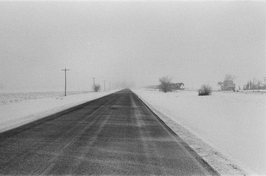 A desolate road through the barren, snowy plains. A train runs parallel to the road, its lights cutting through the fog.