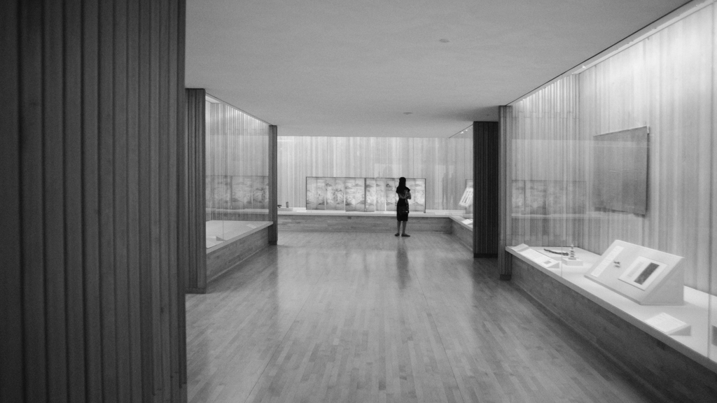 A woman stands alone in a museum gallery. The room has big open displays with only a few objects in each, all behind panes of glass.